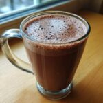 A clear glass mug filled with creamy, frothy hot cocoa mix resting on a wooden surface near a window.