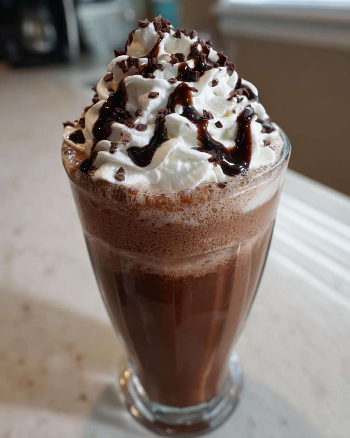 A tall glass filled with rich frozen hot chocolate topped with whipped cream, chocolate syrup, and shavings.