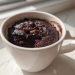 Close-up of a rich, fudgy chocolate mug cake topped with melted chocolate chips, sitting in a white mug.