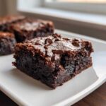 Close-up of a fudgy, dark chocolate sweet potato brownies slice topped with melted chocolate chips on a white plate.