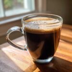 A dark, foamy beverage in a clear glass mug, possibly made from hot cocoa mix, sitting on a sunlit wooden table.