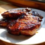 Close-up of two richly glazed, caramelized pork steak recipe cuts resting on a white plate in sunlight.