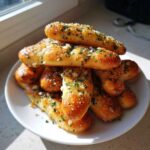 A pile of golden, buttery breadsticks generously topped with garlic and parsley served on a white plate.