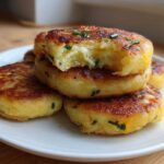 A stack of golden brown, pan-fried mashed potato cakes garnished with chives, with one cake bitten into showing the fluffy interior.