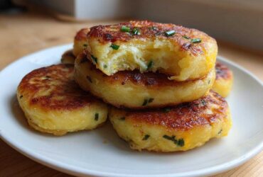 A stack of golden brown, pan-fried mashed potato cakes garnished with chives, with one cake bitten into showing the fluffy interior.