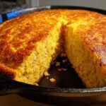 Close-up of golden brown skillet cornbread, showing a slice cut out to reveal the moist interior.
