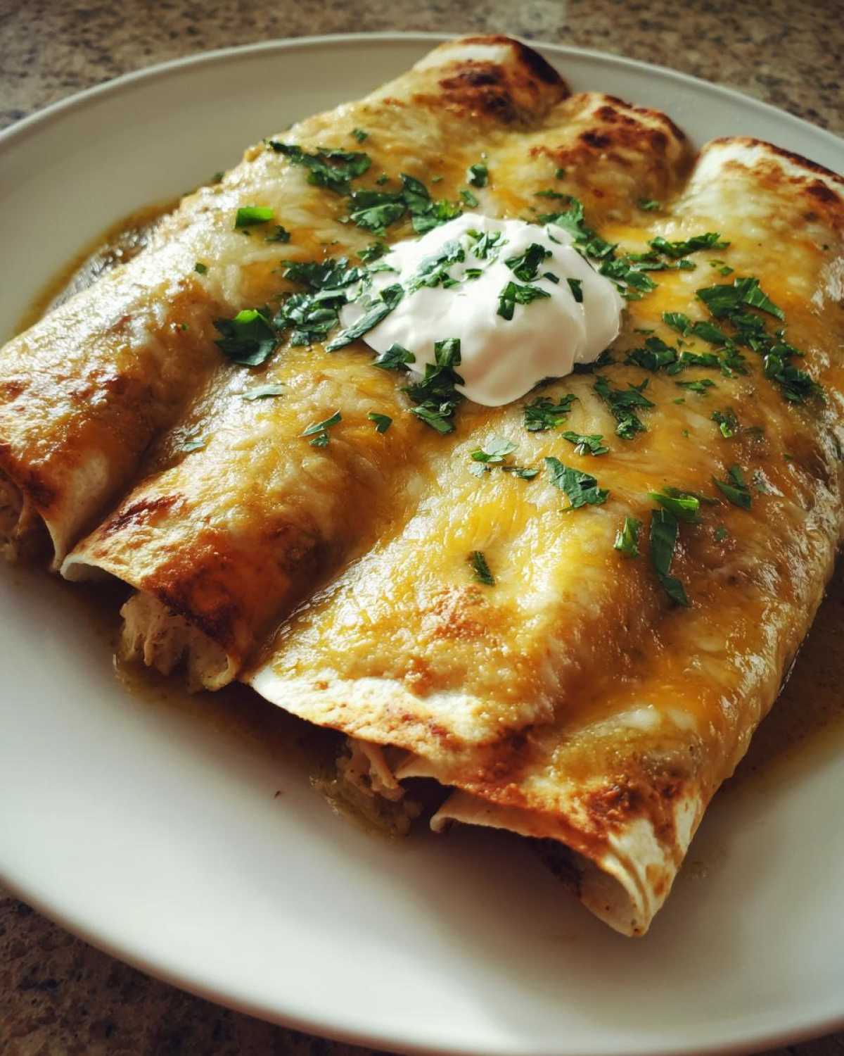 Three delicious green chile chicken enchiladas covered in melted cheese, sour cream, and cilantro on a white plate.