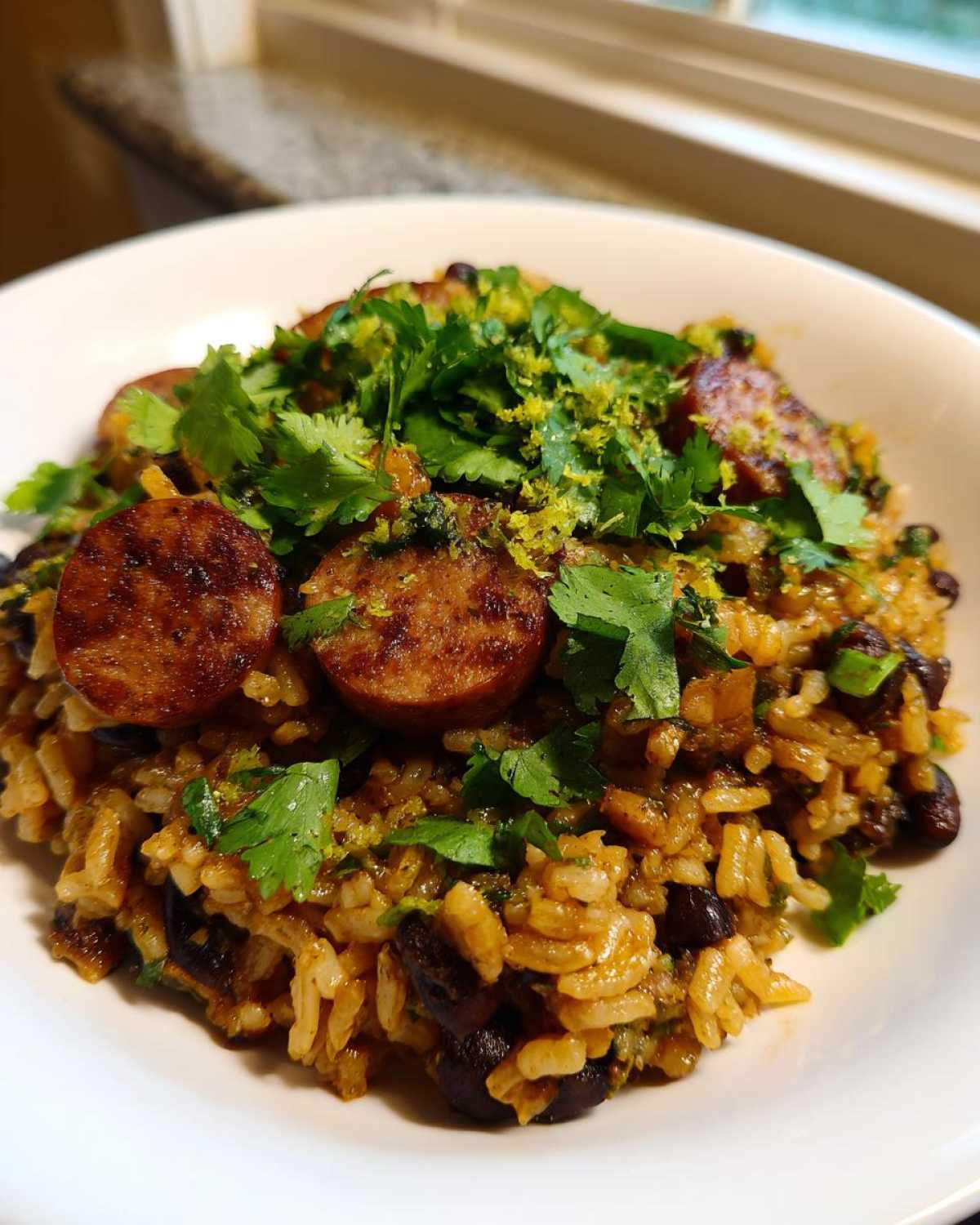 A close-up of a bowl of seasoned black beans and rice topped with sliced, seared sausage and fresh cilantro.