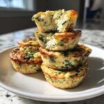 A stack of four golden-brown, high-protein egg bites with visible spinach, one on top broken open.