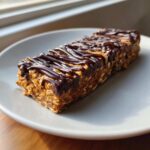 A close-up of a homemade, oat-based protein bar drizzled generously with melted dark chocolate, sitting on a light gray plate.