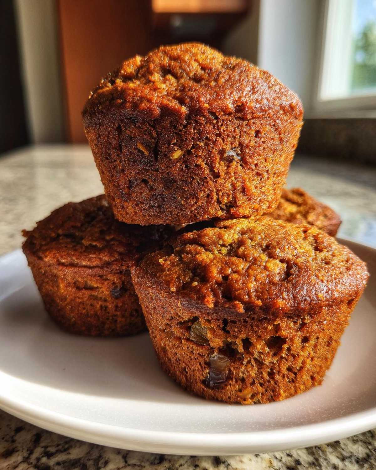 Close-up of three freshly baked, dark brown honey bran muffins stacked on a white plate.