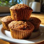 A close-up stack of four freshly baked, dark brown honey bran muffins sitting on a white plate.