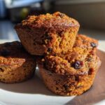 A close-up stack of four golden brown honey bran muffins dotted with raisins, sitting on a white plate.