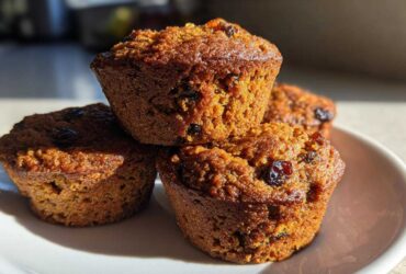 A close-up stack of four golden brown honey bran muffins dotted with raisins, sitting on a white plate.
