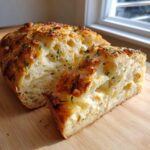 A loaf of Italian herb and cheese bread sliced open on a wooden board, showing a soft, airy interior.