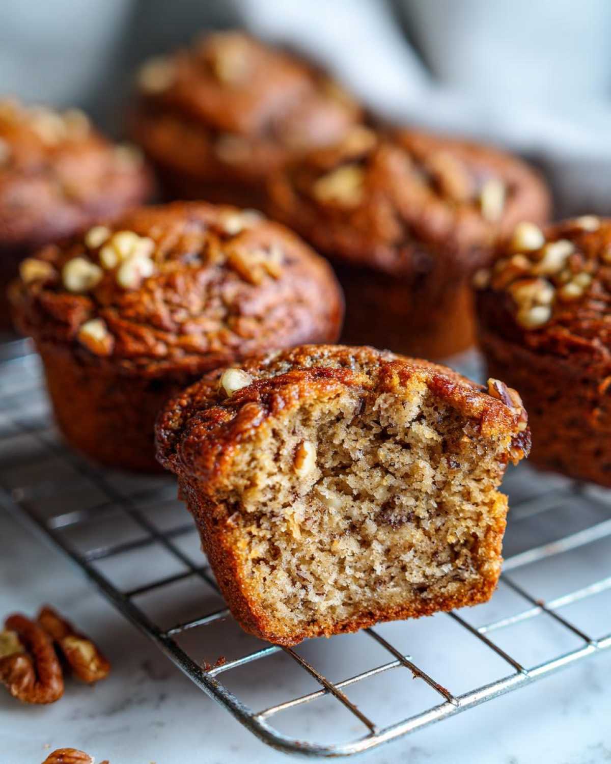 Close-up of a moist banana nut muffin, split open to show the texture, resting on a cooling rack with more muffins behind.