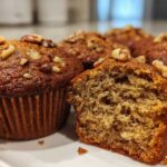 Close-up of moist banana nut muffins, one cut open showing the texture, topped with walnuts.