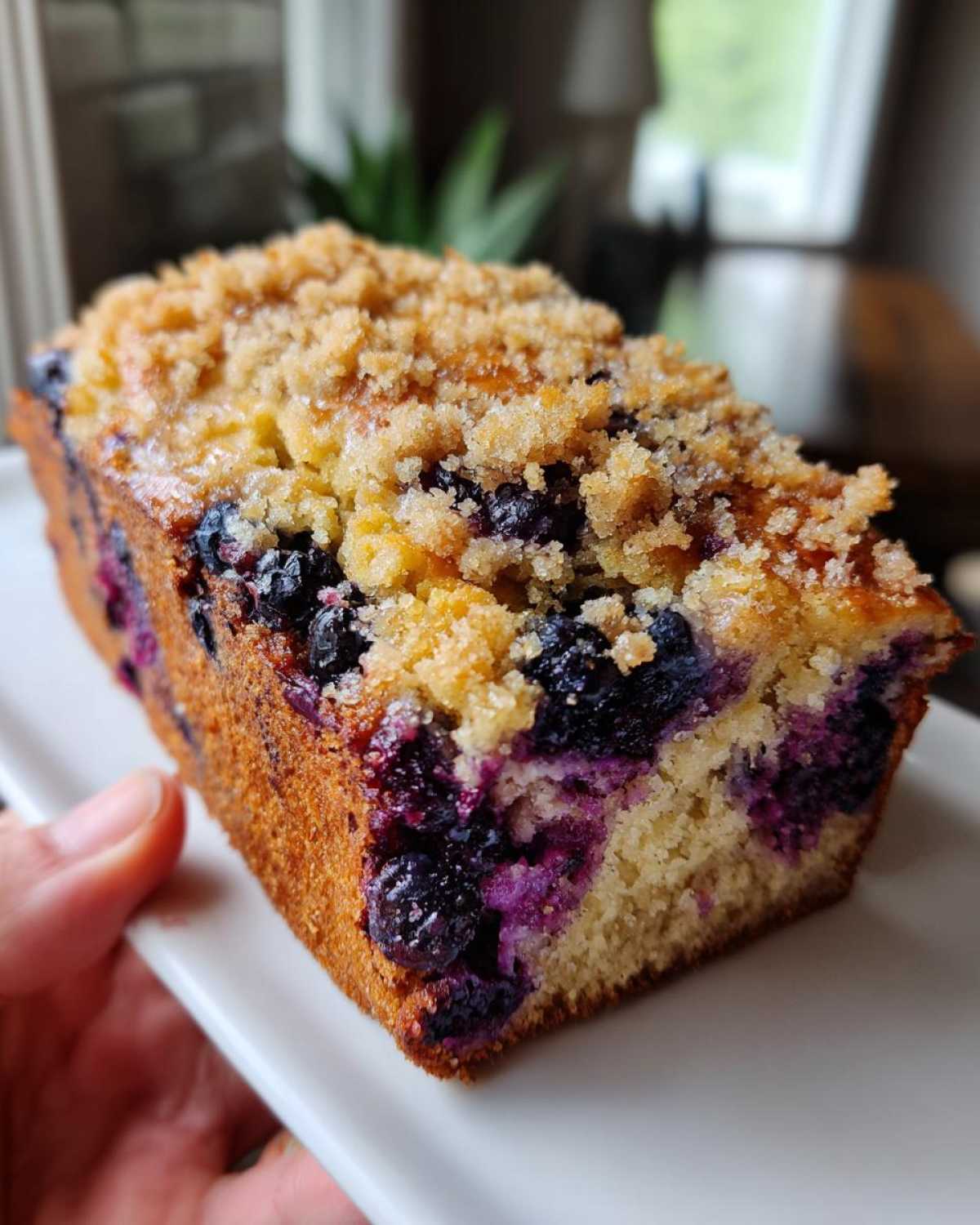 A hand holding a slice of moist blueberry banana bread topped with a sweet, golden crumb topping.