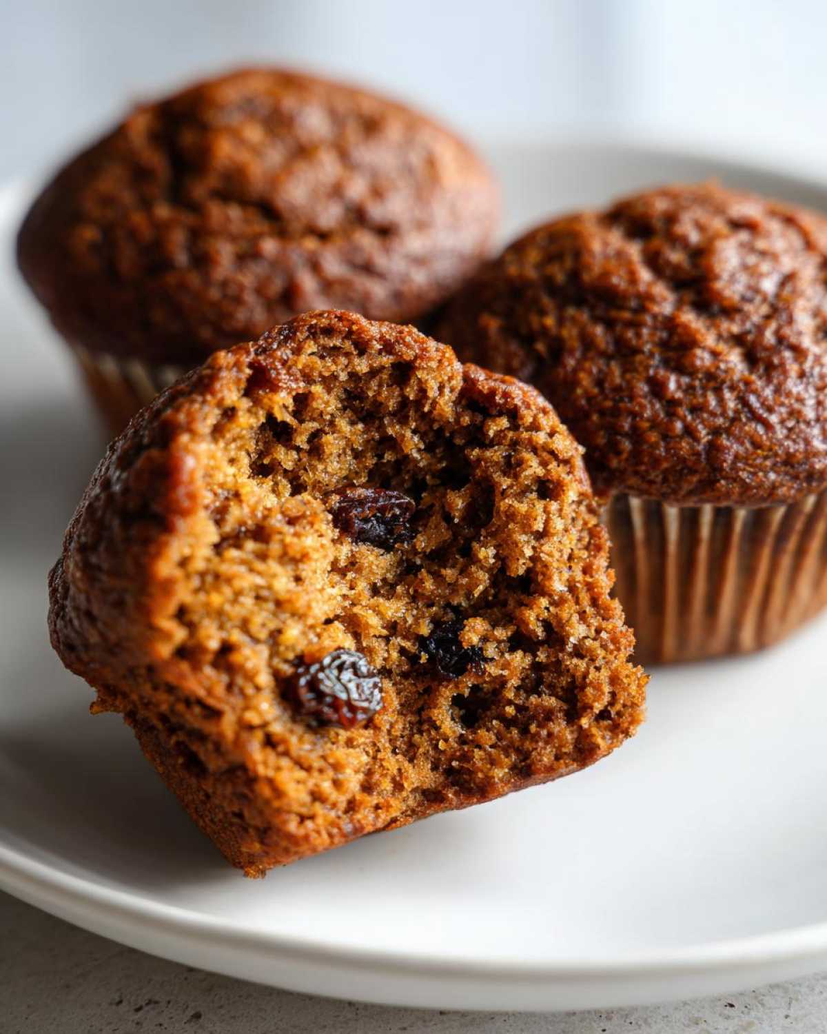 Close-up of a bran muffin cut in half showing the moist texture and raisins inside, with two whole bran muffins blurred in the background.