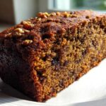 Close-up of a moist slice of dark, rich date nut bread topped with chopped walnuts, sitting on a white plate.