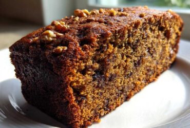 Close-up of a moist slice of dark, rich date nut bread topped with chopped walnuts, sitting on a white plate.