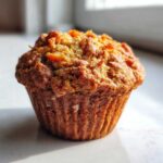 A close-up, appetizing photo of a single, deeply browned morning glory muffin with a textured, slightly craggy top.
