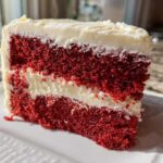 Close-up of a moist slice of red velvet cake showing two vibrant red layers separated by white cream cheese frosting.