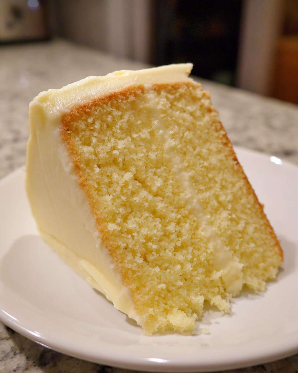 Close-up of a moist slice of vanilla cake recipe with thick, creamy white frosting on a white plate.