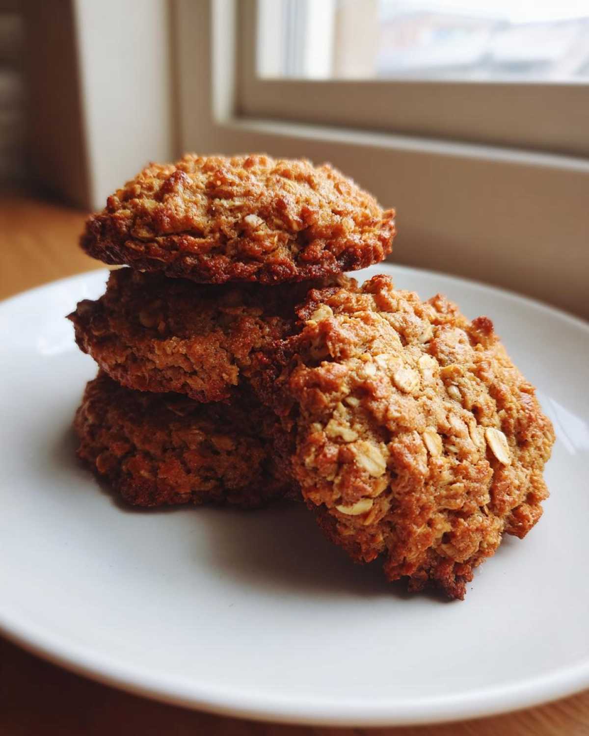 A stack of four golden brown, textured oatmeal cookie treats resting on a white plate near a window.