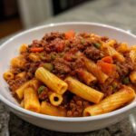 Close-up of rigatoni pasta generously topped with rich, meaty bolognese sauce and visible vegetables.