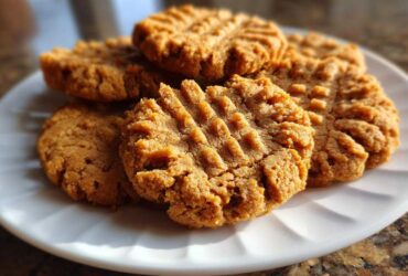 A close-up of several golden brown peanut butter cookies with classic fork marks stacked on a white plate.