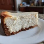 A thick slice of creamy Protein Cheesecake with a browned top edge and dark graham cracker crust on a white plate.