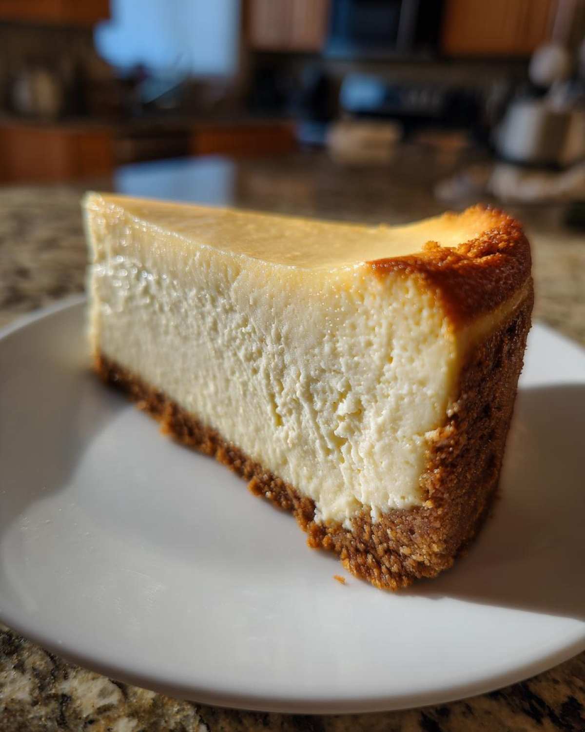 A close-up, sunlit slice of creamy Protein Cheesecake with a thick graham cracker crust on a white plate.