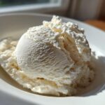 Close-up of a perfectly scooped serving of vanilla-colored protein ice cream in a white bowl, highlighted by sunlight.