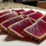 Slices of perfectly seared ahi tuna displaying a deep red center and a thick sesame seed crust on a white plate.