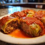 Close-up of several cabbage rolls topped with bright red tomato sauce, one cut open showing the meat and rice filling.