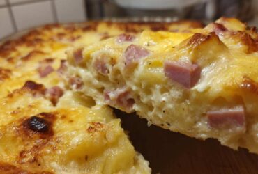 Close-up of a creamy slice being lifted from a baked scalloped potatoes and ham dish, showing melted cheese and diced ham.
