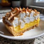 Close-up of a perfect slice of lemon meringue pie showing thick yellow curd and tall, toasted meringue peaks.