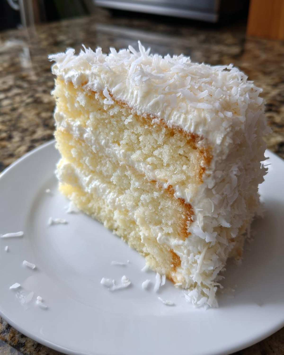 A tall slice of moist coconut cake layered with white frosting and covered in shredded coconut flakes.