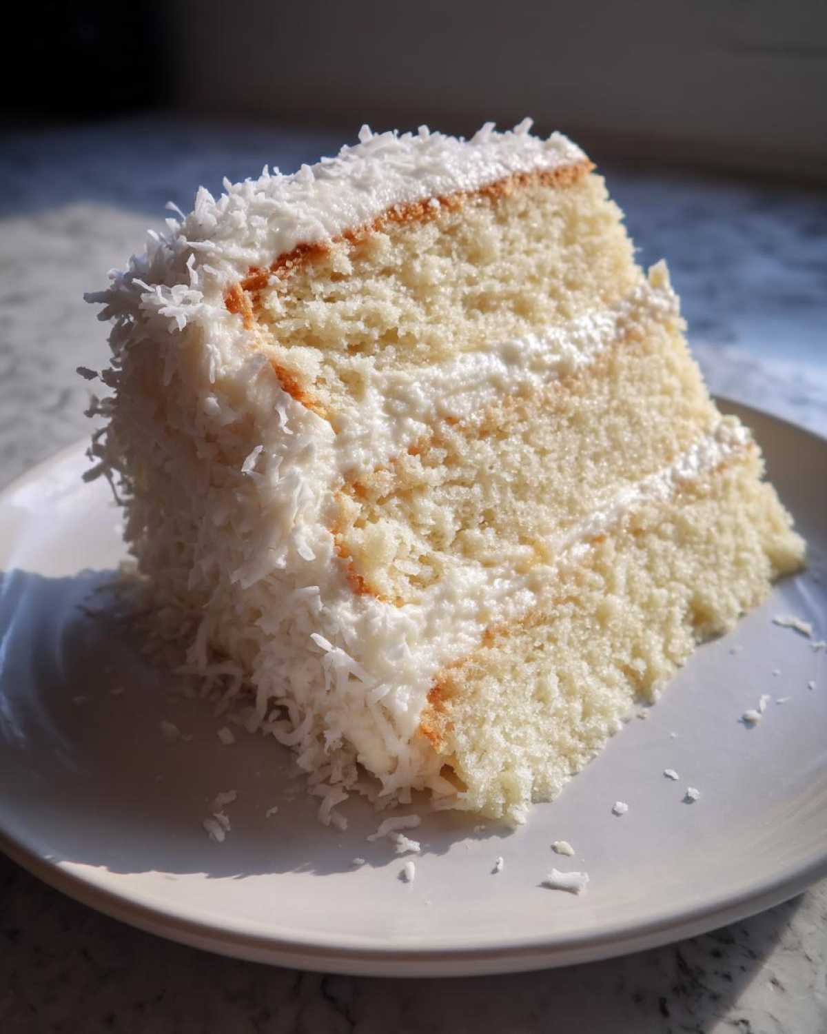 A close-up slice of moist coconut cake layered with frosting and covered in shredded coconut.