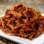 A close-up of saucy, shredded Slow Cooker Pulled Pork piled high on a white rectangular plate.