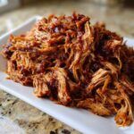 A close-up of shredded, saucy Slow Cooker Pulled Pork piled high on a rectangular white serving dish.