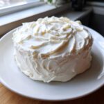 A small, round cake generously covered in thick, swirled cream cheese frosting on a white plate.