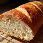 A close-up of a freshly baked loaf of soft French bread, cut open to show the airy crumb, resting on a wire cooling rack.