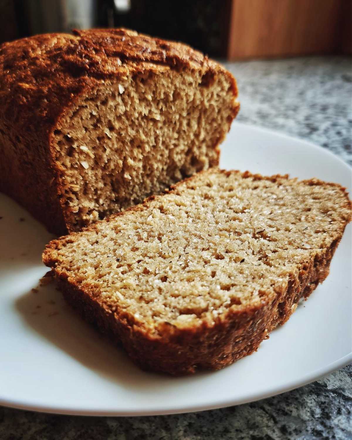 Close-up of a freshly baked loaf of soft whole wheat bread with one thick slice cut and resting beside it on a white plate.