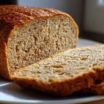 Close-up of a freshly baked loaf of soft whole wheat bread, partially sliced on a white plate.