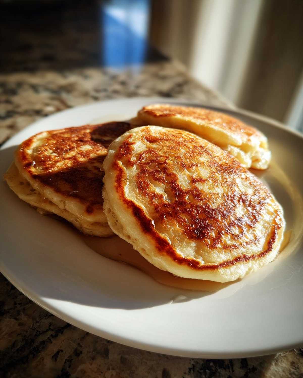 Three fluffy sourdough pancakes drizzled with syrup, sitting on a white plate in bright morning light.