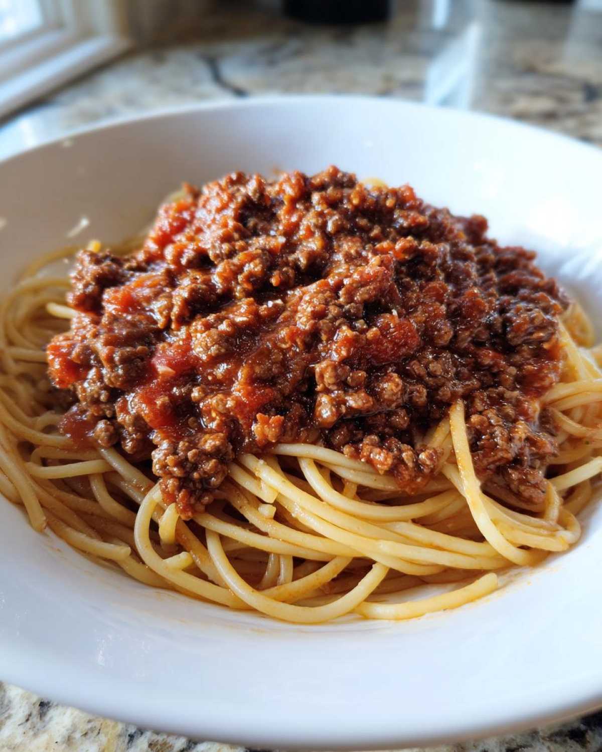 Close-up of spaghetti noodles generously topped with rich, meaty bolognese sauce in a white bowl.