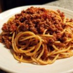 Close-up of spaghetti noodles generously topped with rich, meaty bolognese sauce on a white plate.