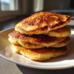 A stack of four fluffy, golden-brown sourdough pancakes on a white plate, highlighted by bright sunlight.